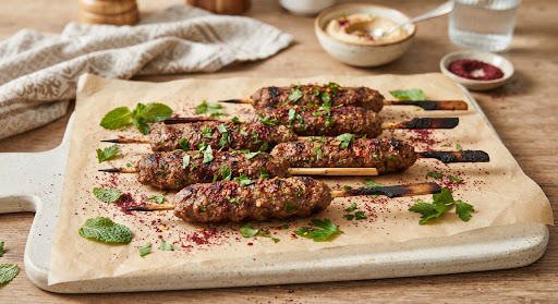 Six artisanal seasoned lamb kofta skewers on wooden skewers arranged neatly on parchment paper with sumac and fresh mint…